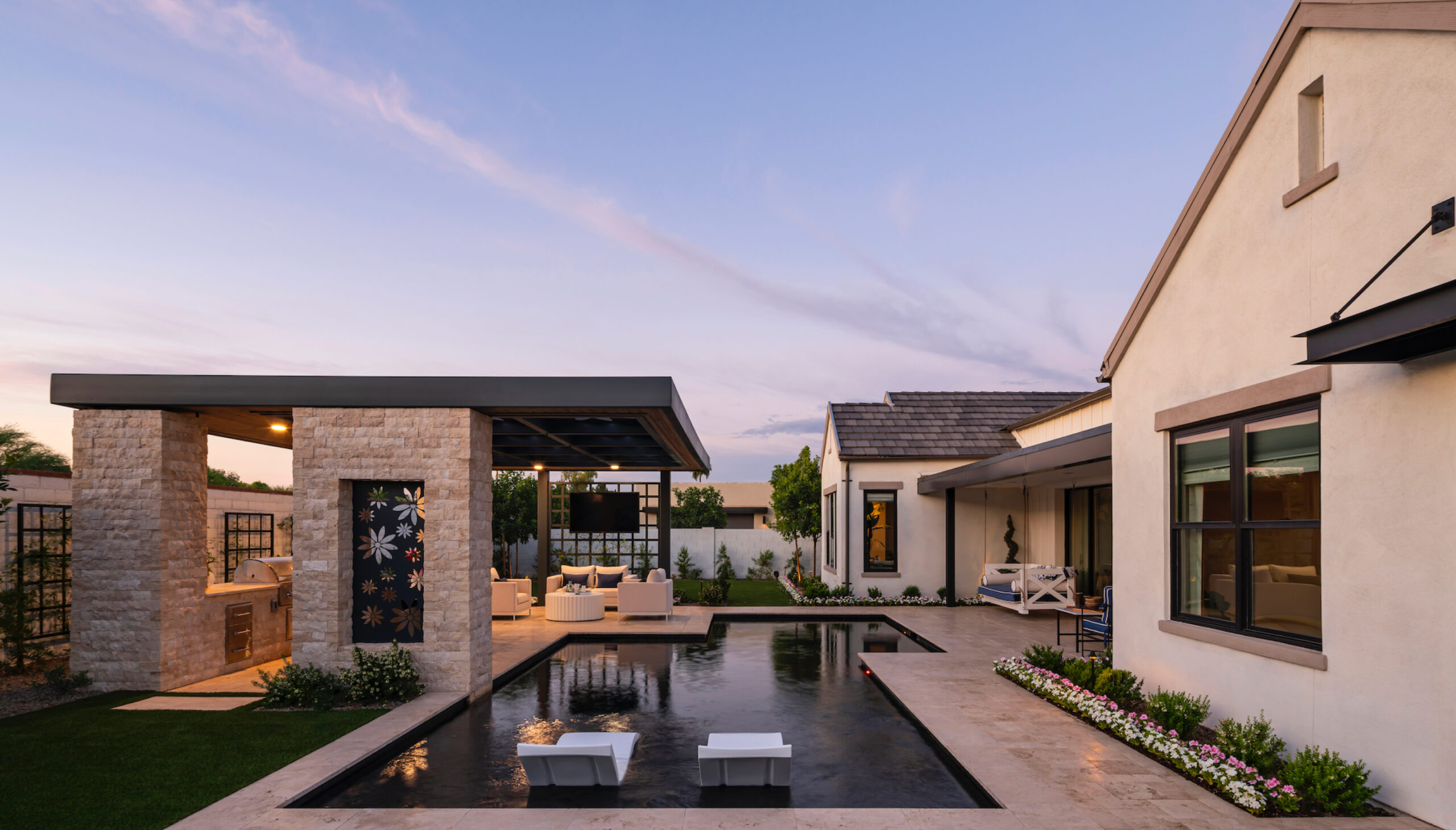 Modern house with a poolside patio, outdoor seating, and a pergola. The sky is clear, and the garden is landscaped with small plants and flowers.